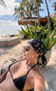 Woman in a black bikini and headscarf sitting on a beach with palm trees and a hut in the background.